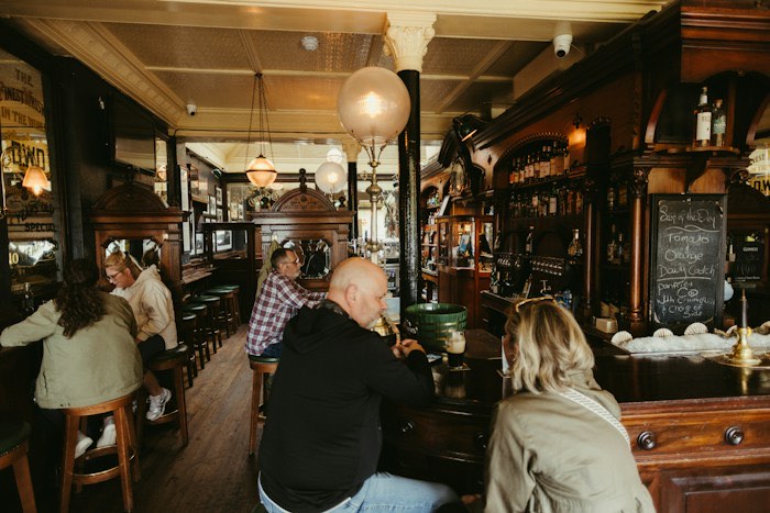 Gjester ved bardisken og bordene i en tradisjonell pub med mørkt treverk og glasskupler i taket